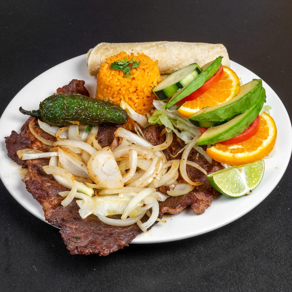 Grilled steak topped with onions, orange rice, burrito, roasted pepper, avocado, tomatoes, citrus slices, and lime on a white plate.