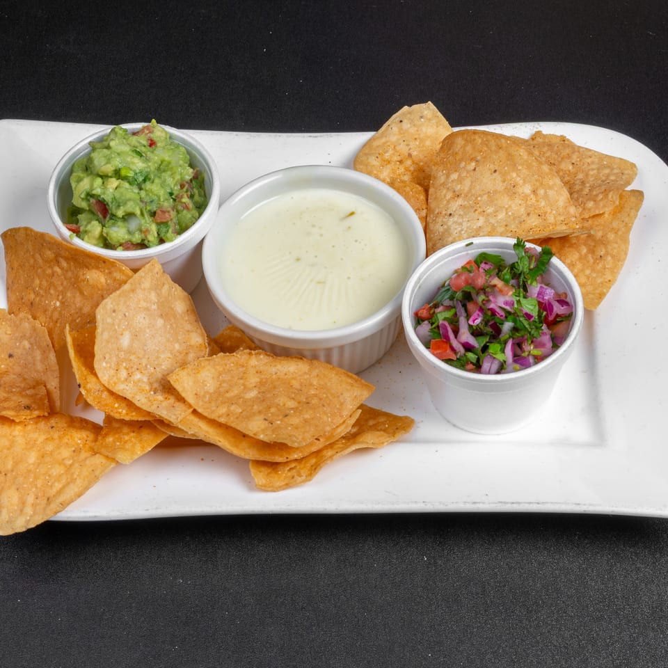 Crispy tortilla chips served with guacamole, creamy white dip, and fresh tomato-onion salsa on a white platter.