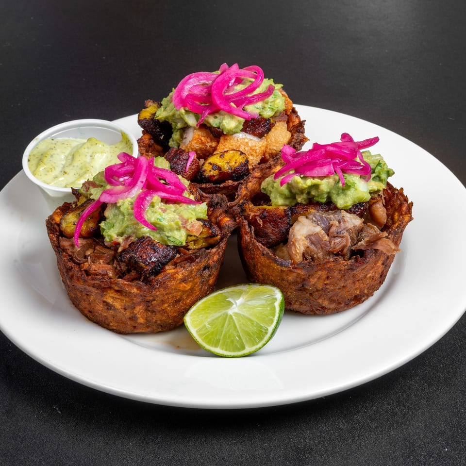 Three crispy plantain cups filled with meat, fried plantains, guacamole, and pickled onions, served with sauce and lime.
