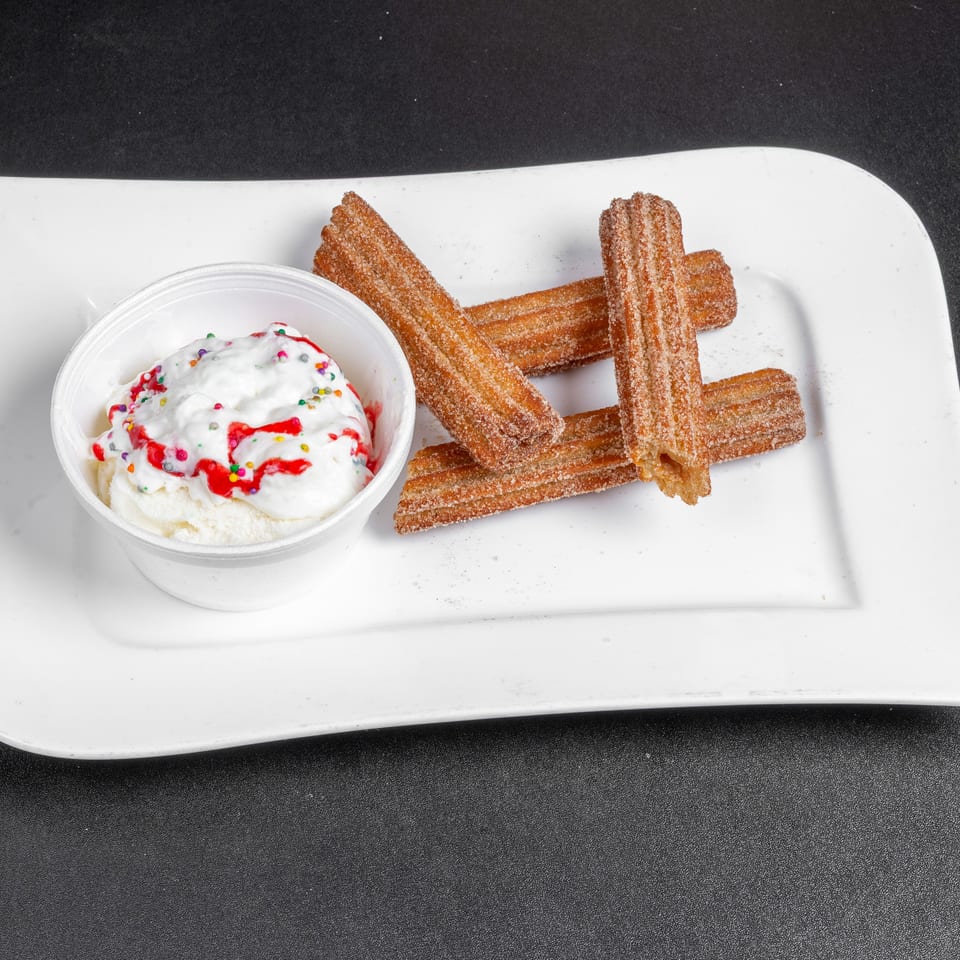 Cinnamon-sugar churros served with whipped cream, red syrup, and colorful sprinkles on a white plate.