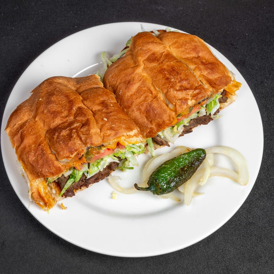 Grilled steak sandwiches with lettuce, tomatoes, onions, and melted cheese on a white plate, served with grilled pepper and onions.