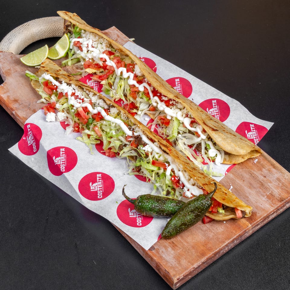 Three loaded tacos with lettuce, diced tomatoes, cotija cheese, crema, lime wedges, and roasted peppers on a wooden board.