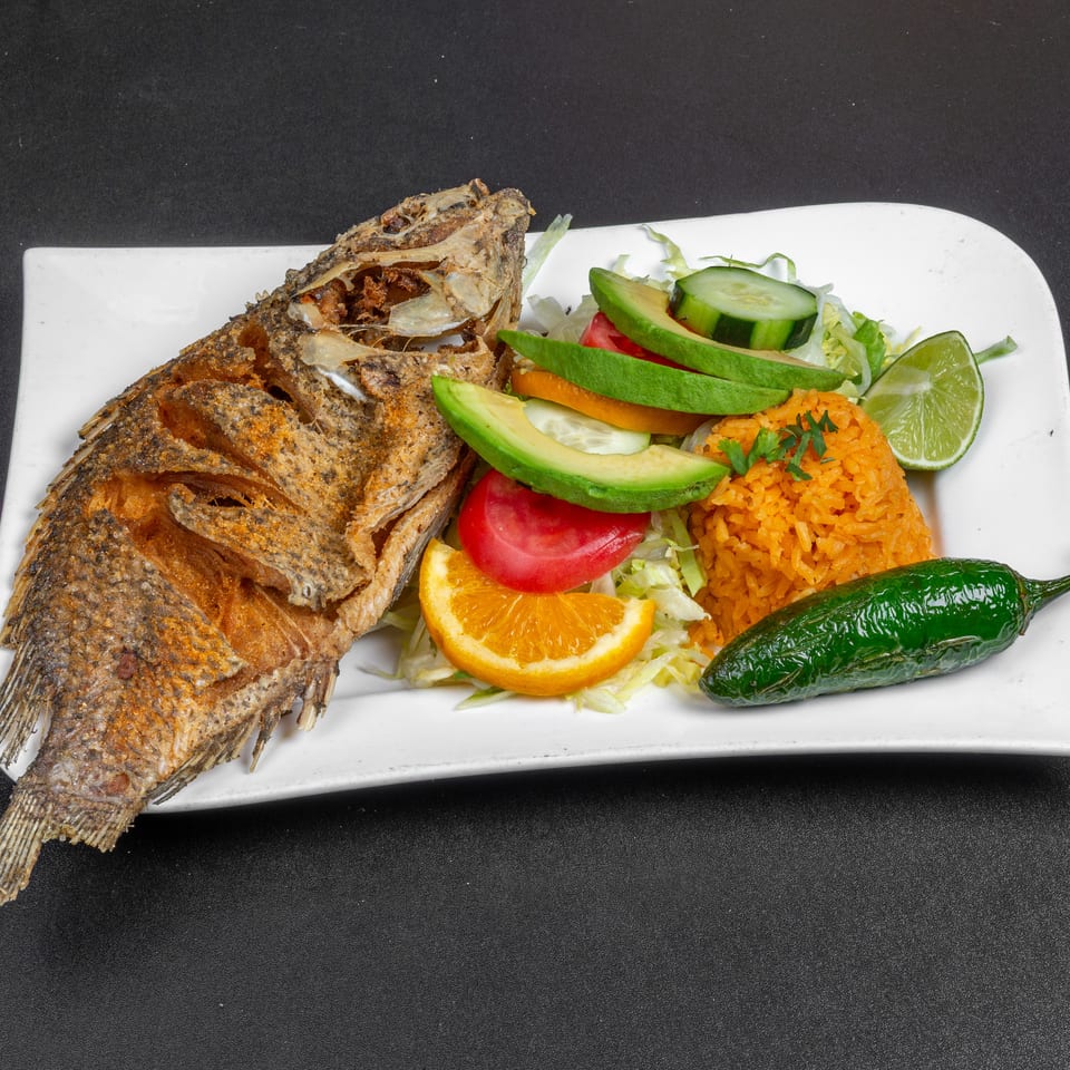 Whole fried fish served with Mexican rice, avocado, salad vegetables, lime, orange, and grilled pepper on a white plate.