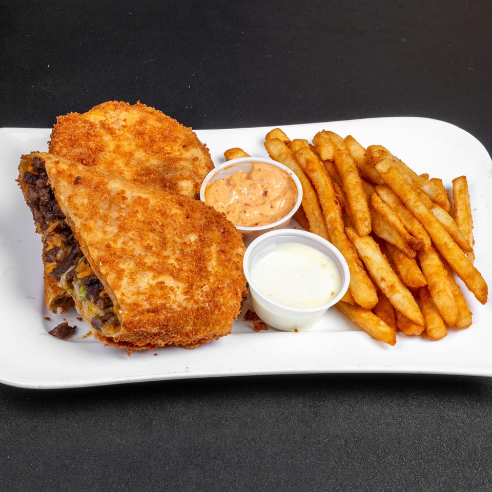 Golden-brown breaded steak sandwich with fries, spicy dipping sauce, and creamy white sauce on a rectangular white plate.