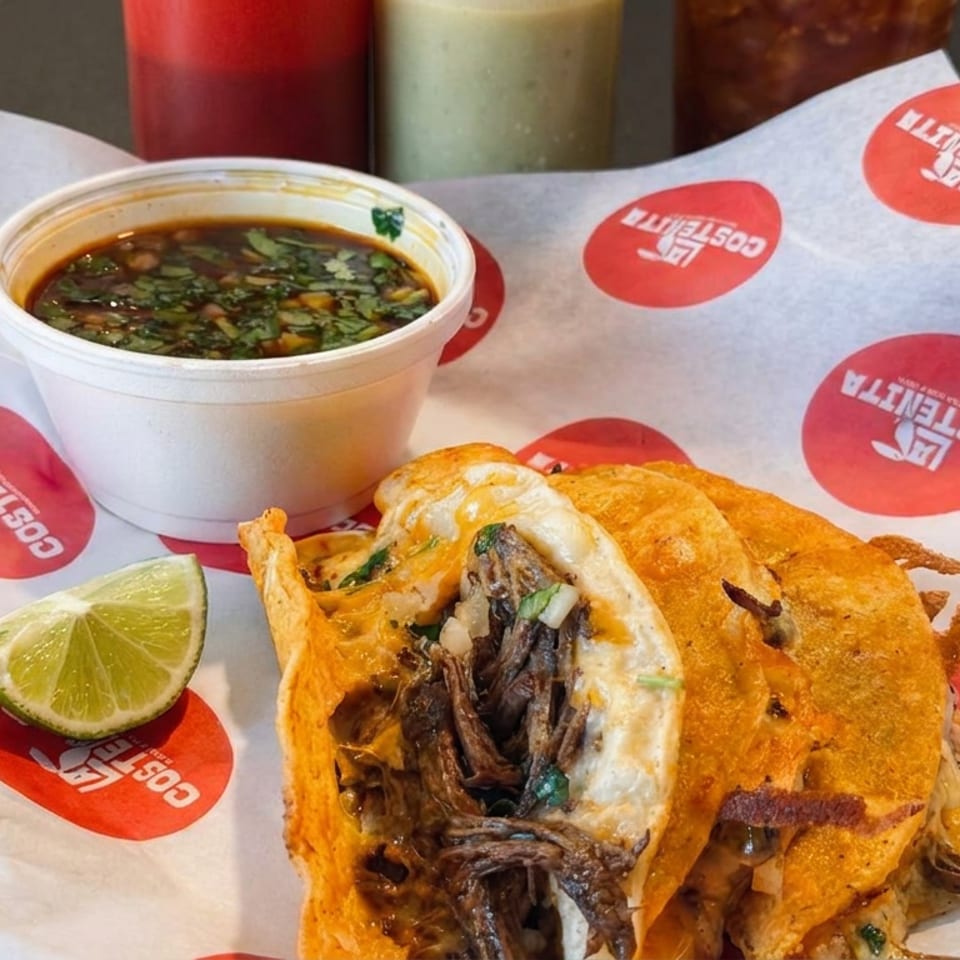 Crispy birria tacos filled with shredded beef beside a cup of consommé and lime wedge on branded paper.
