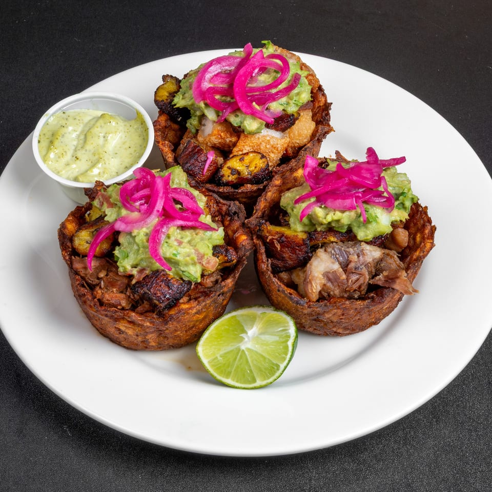 Three crispy plantain cups filled with savory meat, guacamole, and pickled onions, served with dipping sauce and lime.