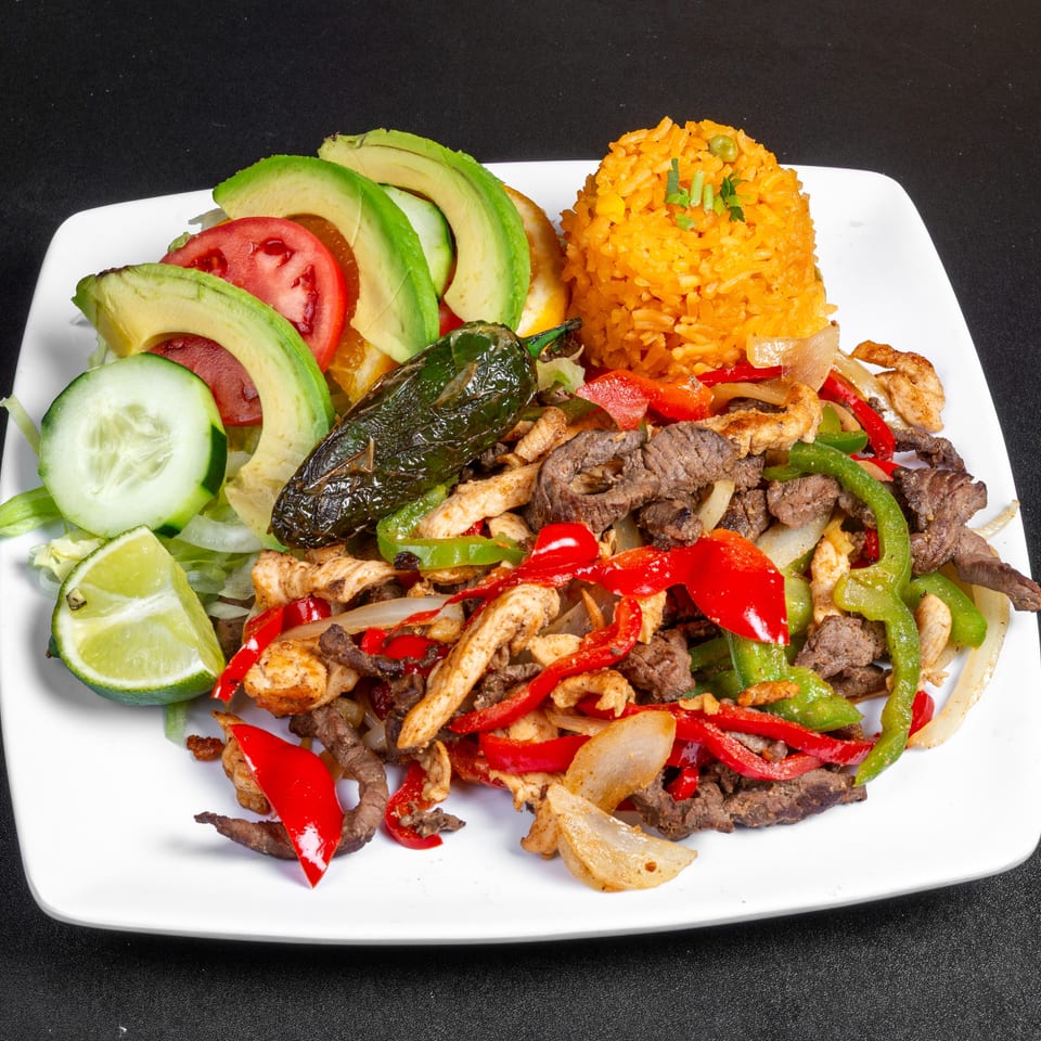 Mixed beef and chicken fajitas with peppers, onions, avocado, vegetables, lime, and orange rice on a white plate.