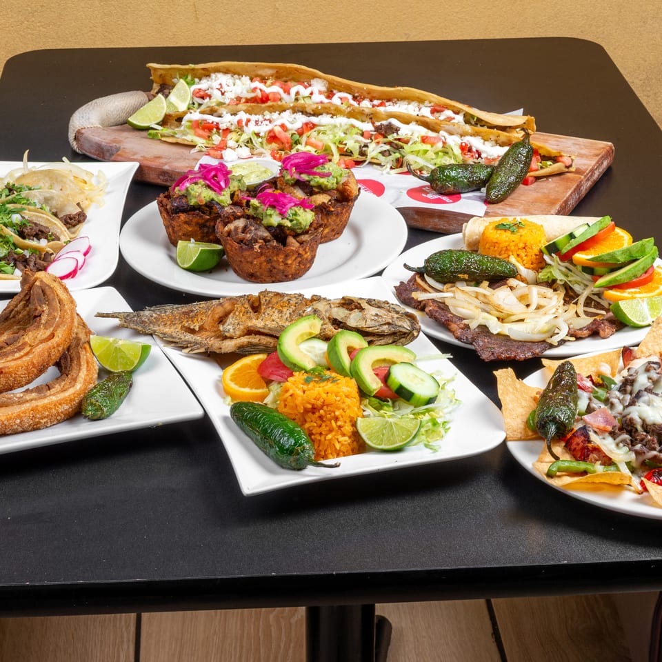 Assortment of Mexican dishes including tacos, enchiladas, grilled meats, fried fish, nachos, rice, guacamole, and roasted peppers.