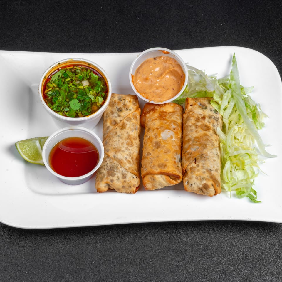 Three crispy fried rolls served with shredded lettuce, lime, cilantro salsa, creamy orange sauce, and sweet chili sauce on a white platter.